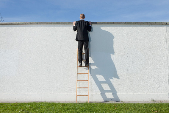 A man in a black suit climbs a wooden ladder to look over a tall white wall on a sunny day. His shadow is cast sharply on the wall, and green grass covers the ground below.
