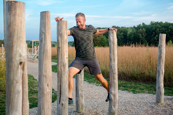 A man exercises outdoors by stepping between tall wooden posts on a gravel path. He grips the posts for balance while moving forward, with grassy fields and trees in the background under a clear sky.