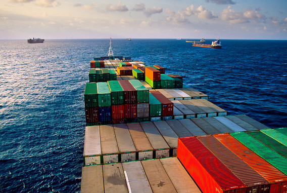 A cargo ship loaded with colorful shipping containers sailing on the open sea under a partly cloudy sky. Several other cargo ships are visible in the distance on the horizon.