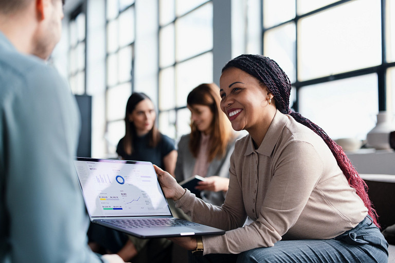 A woman sitting in an office environment, holding a laptop with a financial presentation on the screen. She is smiling at a male colleague sitting in front of her. Two other women are sitting behind her, talking to each other
