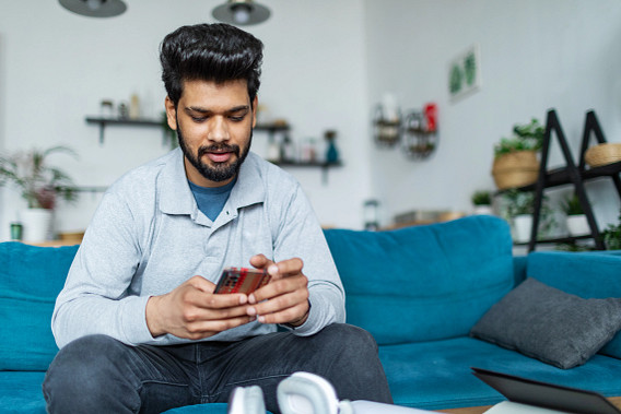 A man sitting on a blue couch in a modern living room, holding a smartphone and looking at it intently for stock trading. The room is decorated with plants, shelves, and a cozy ambiance. A pair of headphones and other items are visible on the table in front of him.