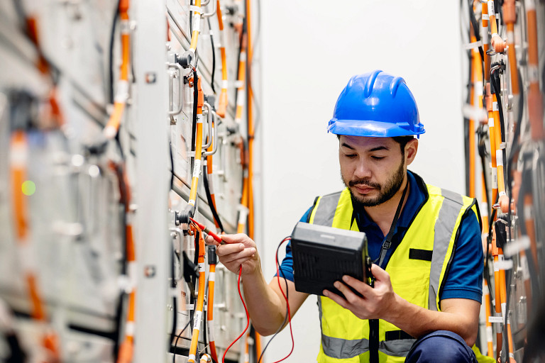 A technician wearing a blue hard hat and high-visibility vest inspects energy storage equipment using a handheld device. The equipment features numerous orange and black cables connected to battery modules in a clean, organized environment.