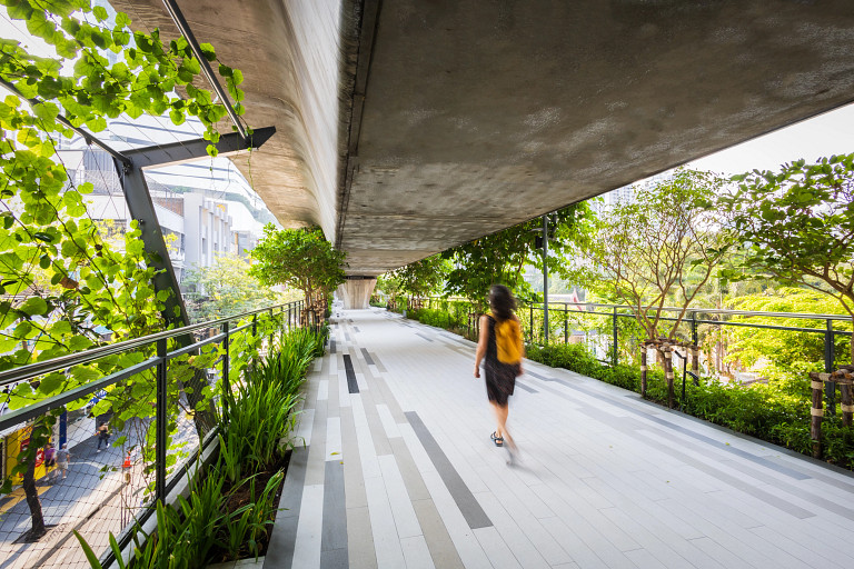 Woman wearing black dress and orange backpack walking on the pedestrian walkway ( Sky Walk) Sukhumvit Line in Bangkok There are green trees on either side of her.
