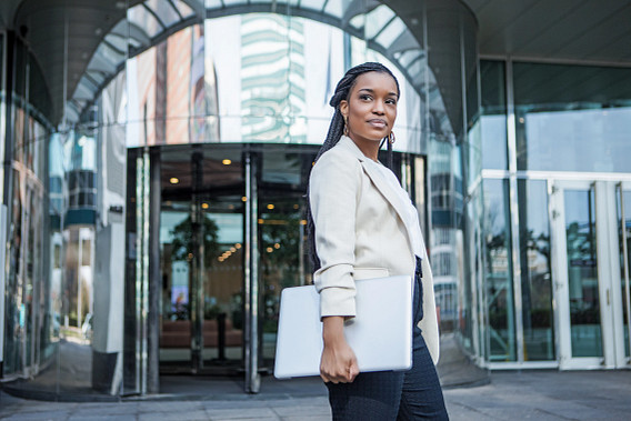 Business woman in light blazer holding laptop outside modern glass office building with revolving doors, reflecting urban surroundings.