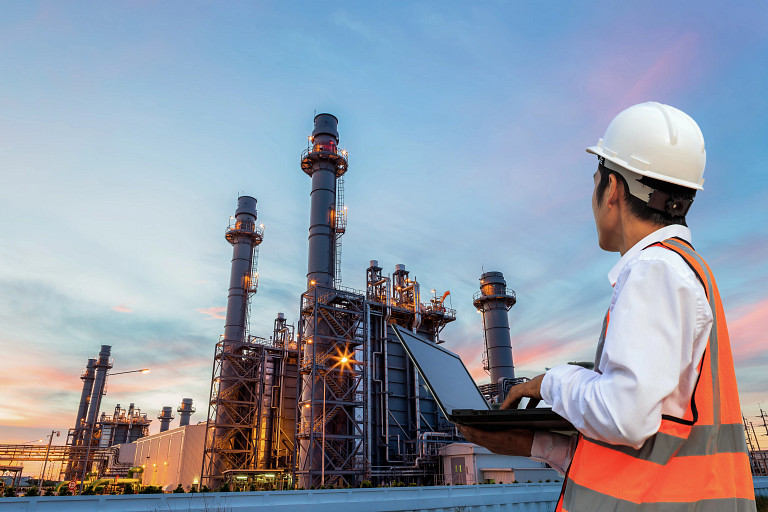An engineer wearing a white helmet and orange safety vest uses a laptop while observing an industrial plant with tall structures and chimneys at sunset.