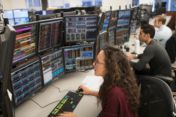 A female in burgundy, a male in dark grey and a male in blue shirt at their trading desk.
