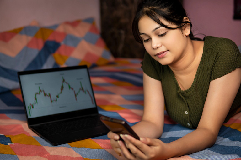 An Indian woman lying on a bed, looking at her smartphone. Behind her, a laptop displays a financial chart with upward and downward trends. The bed has a colorful, patterned blanket, and the setting appears to be a cozy indoor environment.